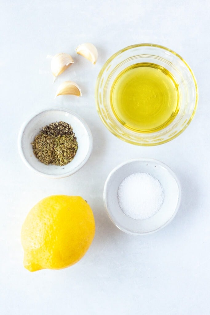 Top down shot of ingredients for lemon chicken marinade on a white background. Includes a lemon, small bowls with herbs, kosher salt, and oil in them, and garlic cloves.