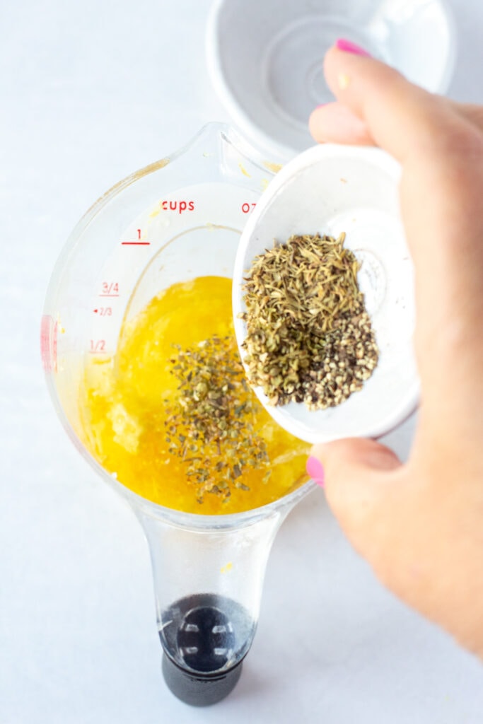 A hand pouring herbs from a small white bowl into a measuring cup with oil and garlic in it.