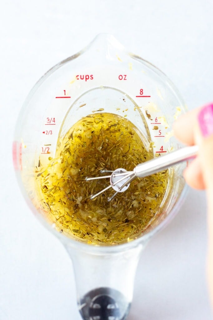 A hand stirring oil, herbs, lemon juice, and garlic together with a small whisk in a measuring cup that rests on a white background.