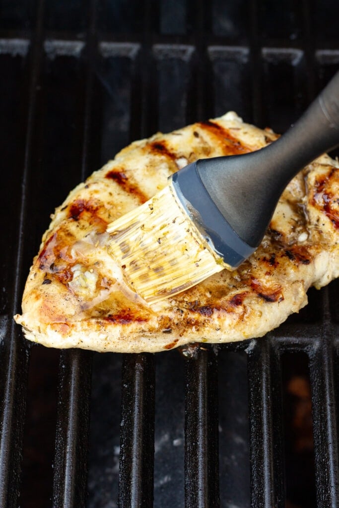 Close up of a basting brush sweeping a lemon glaze on top of a chicken breast cooking on the grill.