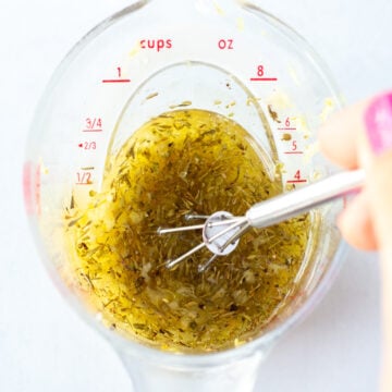 A hand stirring oil, salt, lemon juice, zest, and herbs together in a liquid measuring cup.