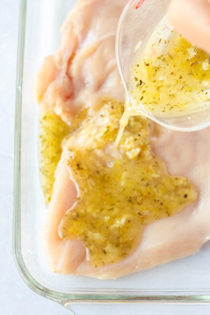 Pouring lemon marinade onto raw chicken breasts in a glass dish.