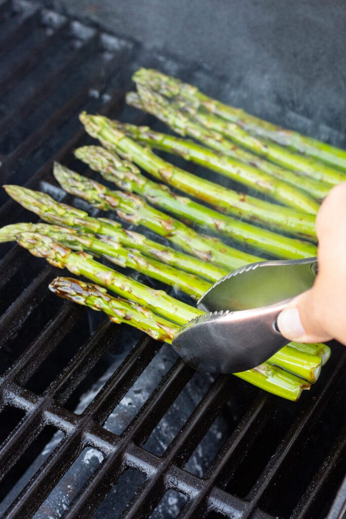 Tongs placing marinated asparagus on a hot grill.
