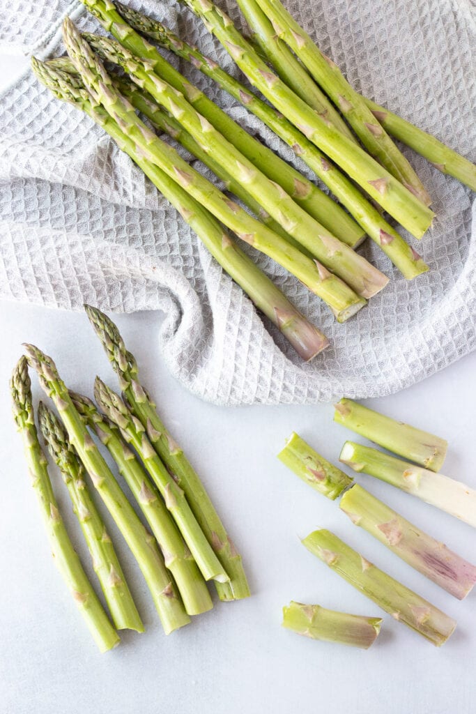 Green asparagus in the process of having their bottoms snapped off, laying on a gray towel and on a white background.