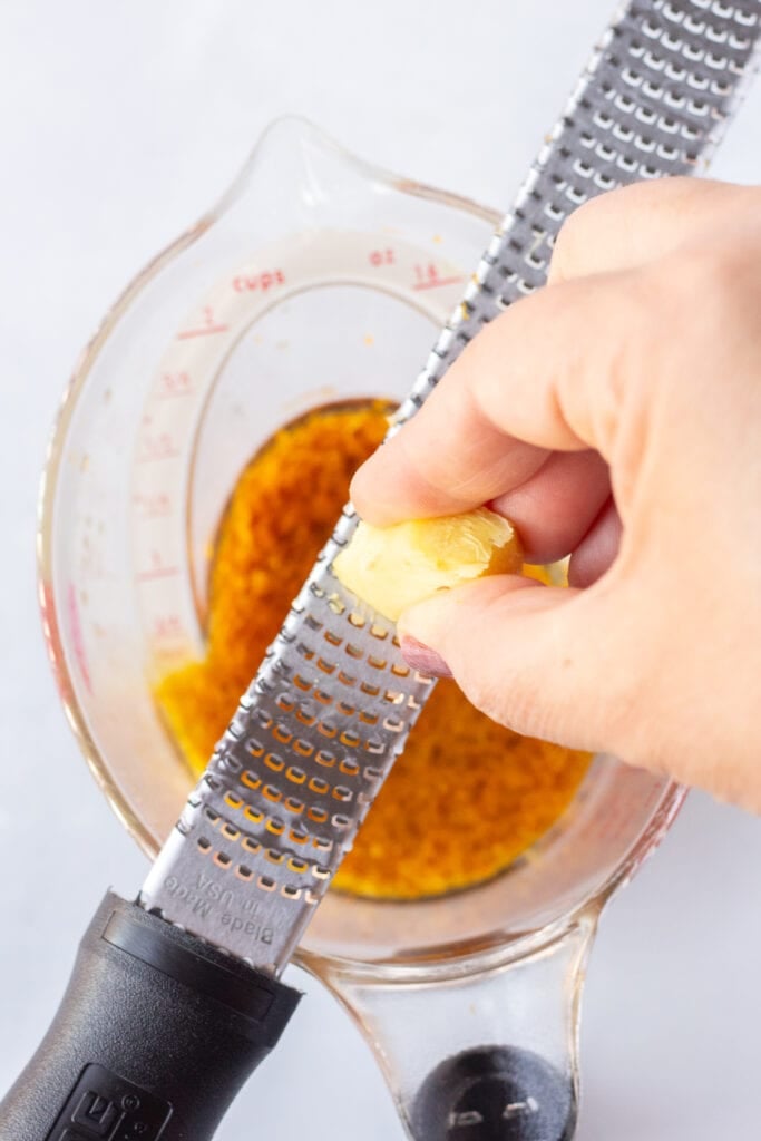 grating a piece of fresh ginger on a microplane over a large liquid measuring cup.