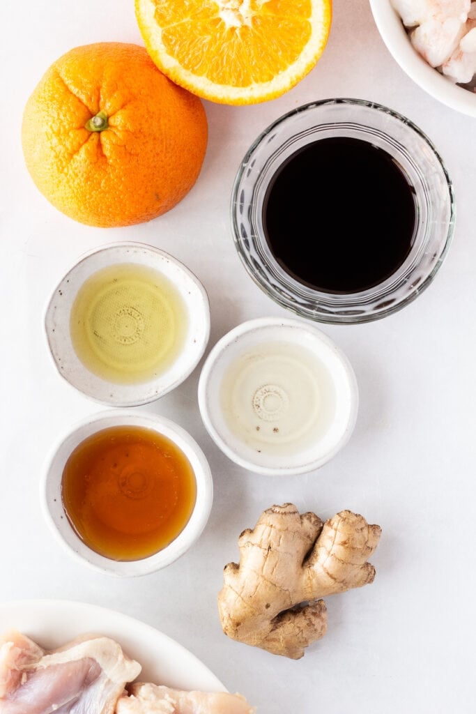 Top down shots of ingredients for orange juice marinade in small bowls, including an orange cut in half, a piece of ginger roote, and honey, soy sauce, oil, and vinegar.