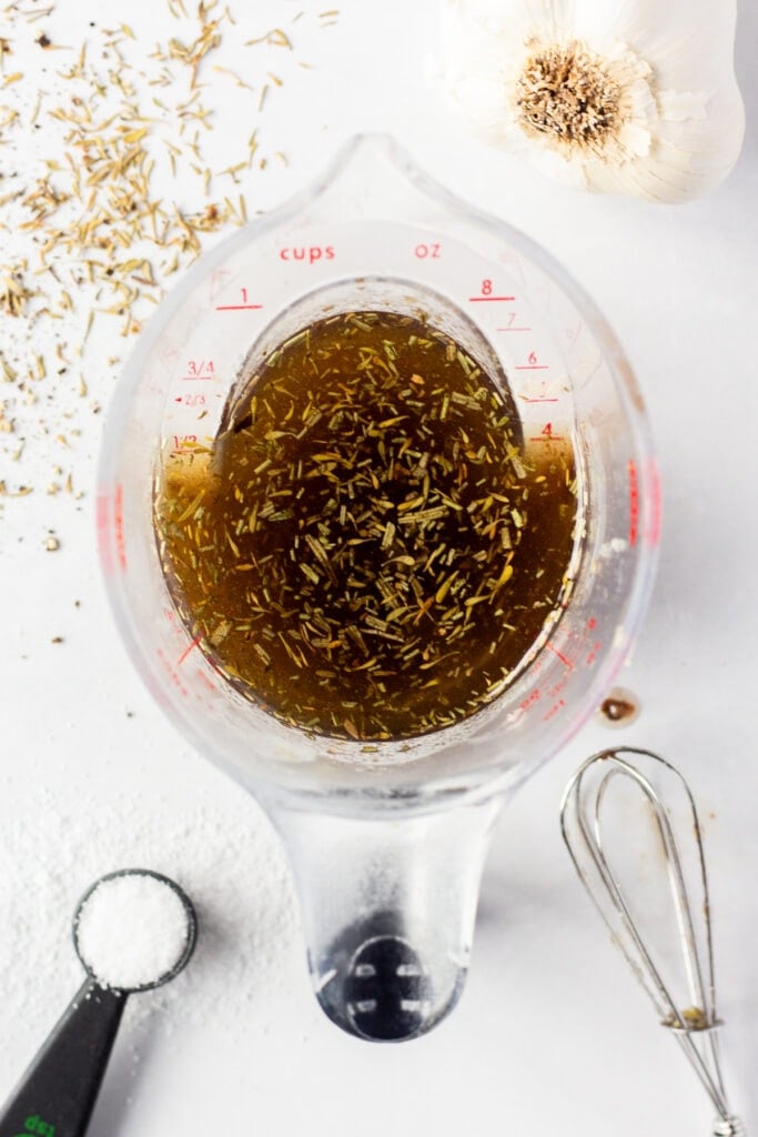 Top down shot of balsamic marinade in a measuring cup with a small whisk, measuring spoon, head of garlic, and dried herbs surrounding it.