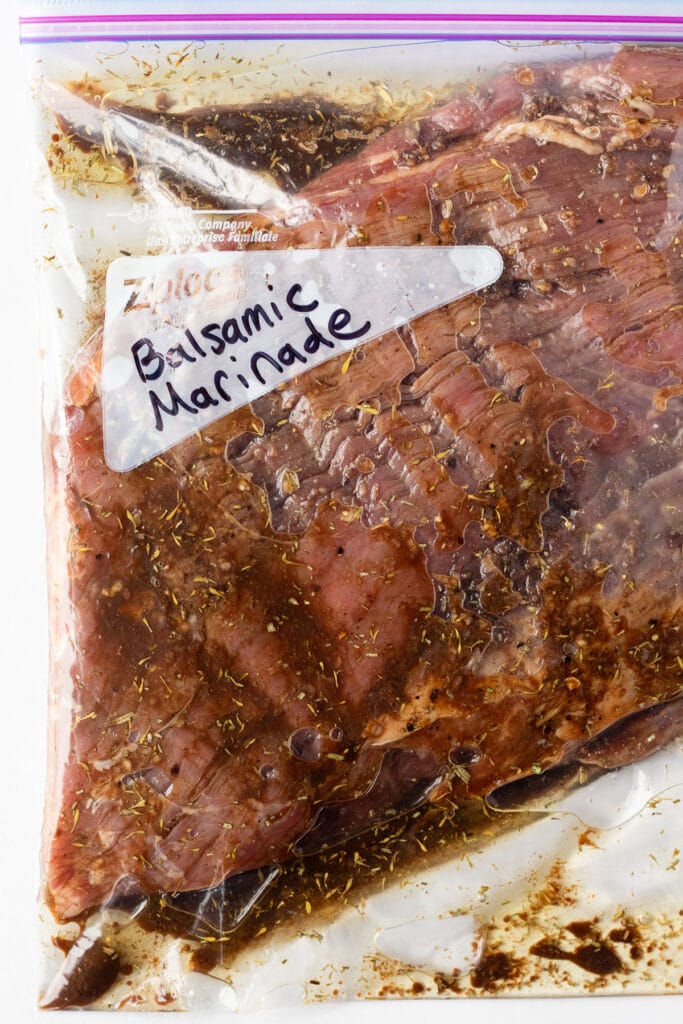A flank steak in a plastic ziploc bag with a balsamic marinade in it, and the words "balsamic marinade" written in black market on the bag.