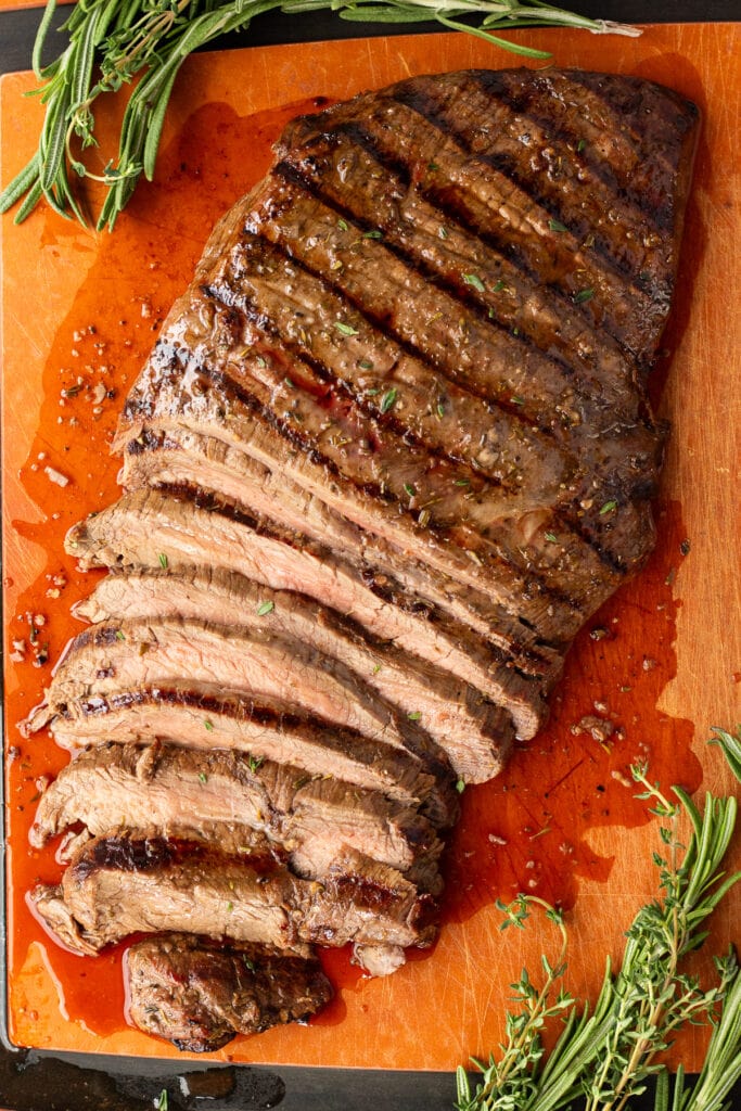 Top down shot of a grilled marinated flank steak on a wood cutting board. Half of it is sliced and there are fresh herbs surrounding it.