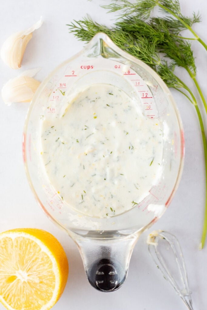 Top down shot of Greek yogurt marinade in a plastic measuring cup on top of a white background with fresh dill, garlic, and half a lemon surrounding it.