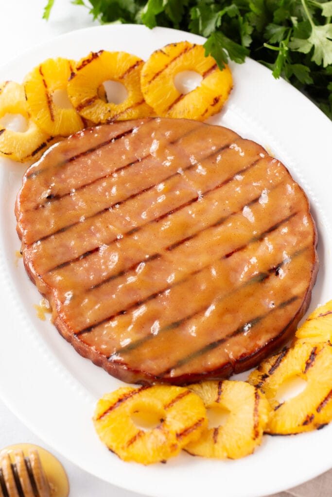 A grilled ham steak with grilled pineapple rings on a white oval platter.
