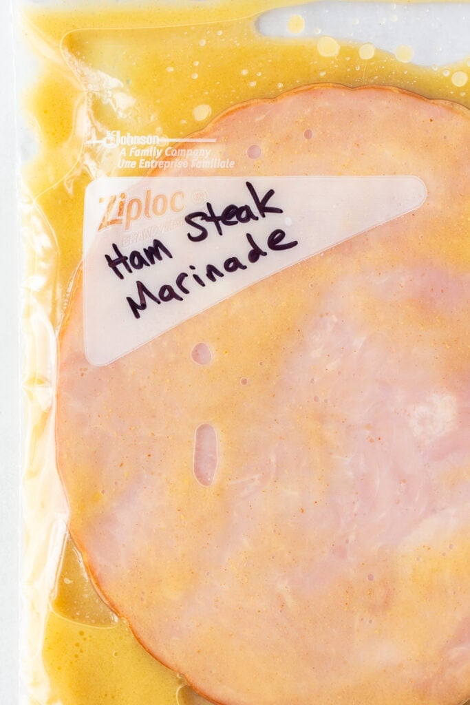 Close up of a ham in a ziploc bag with a honey mustard marinade in it. The words "ham steak marinade" are written in black sharpie on the front.