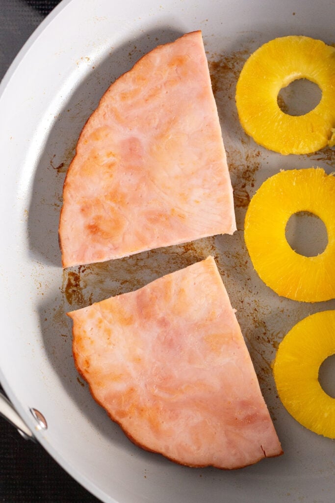Half a ham steak, cut into two pieces, in a large gray pan with pineapple rings beside it.