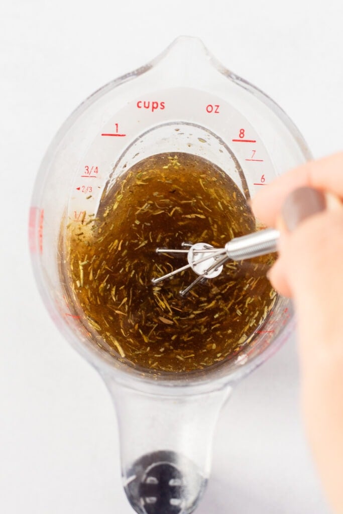 A hand using a small whisk to stir oil, vinegar, and spices together in a clear measuring cup.