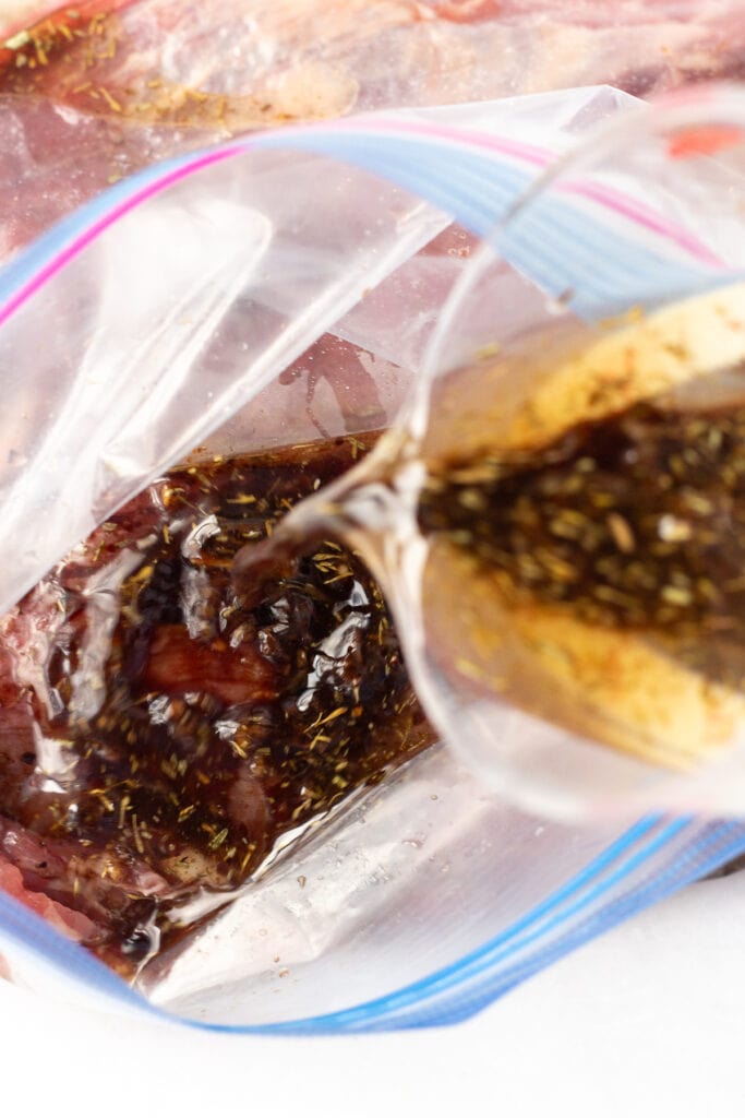 Pouring balsamic marinade from a cup into a ziploc plastic bag with raw flank steak in it.