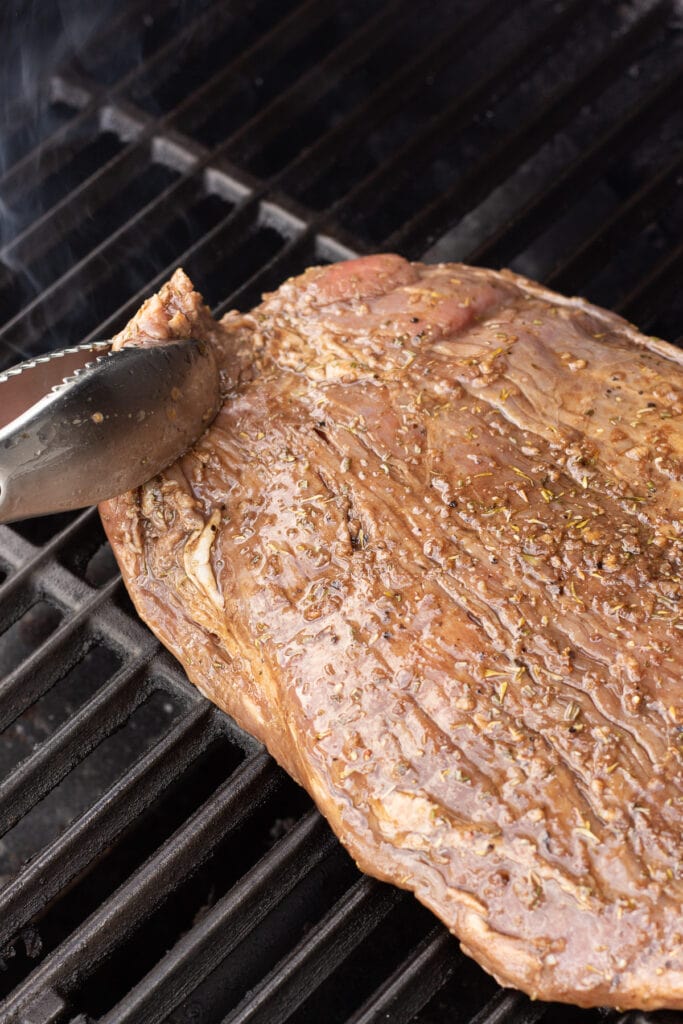 A pair of tongs placing a raw marinated flank steak on a hot grill.