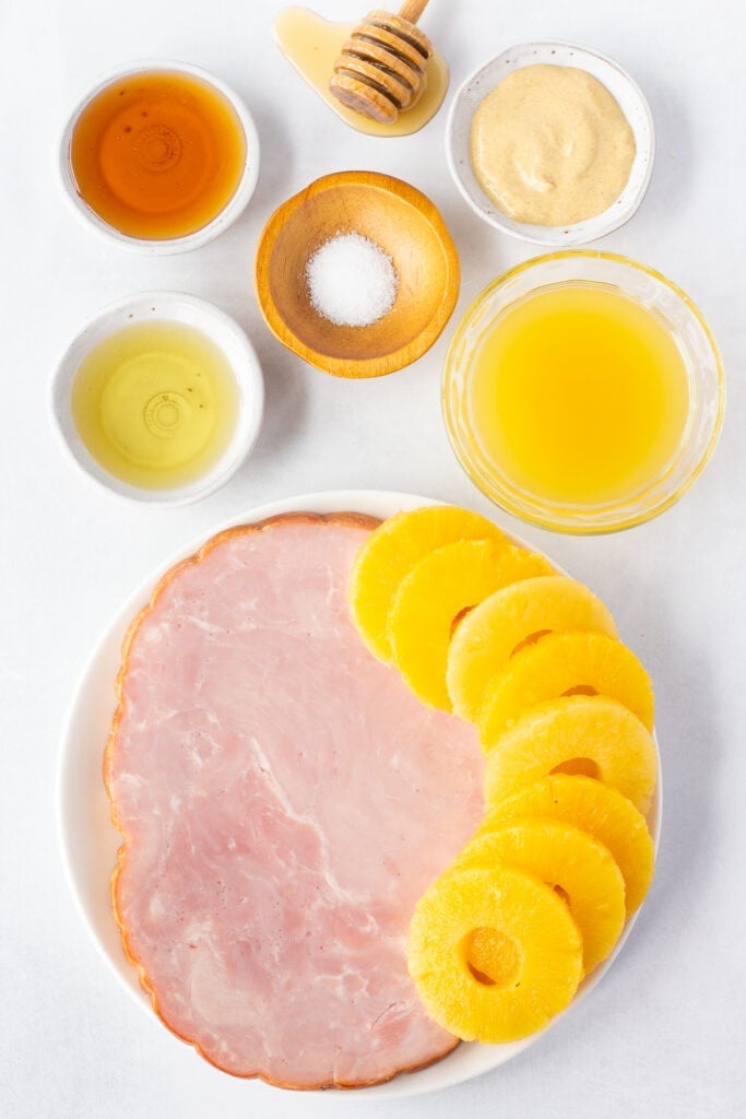 top down shot of ingredients for grilled ham and pineapple on plates and bowls on a light gray background.
