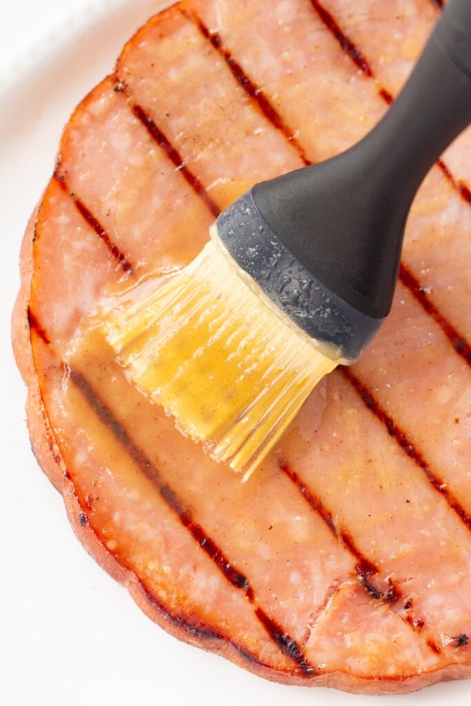 A basting brush wiping glaze onto a grilled ham steak that's on a white platter.