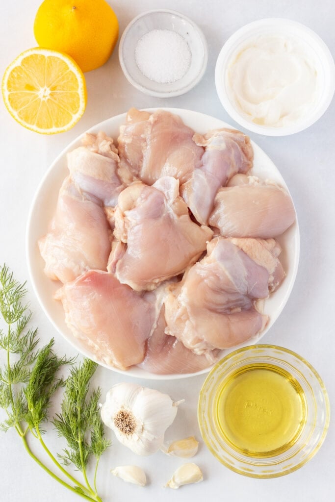 Ingredients for yogurt-marinated Greek chicken thighs on plates and in bowls on a white background. Includes a plate of boneless skinless chicken thighs, a lemon cut in half, oil, plain Greek yogurt, salt, garlic, and dill fronds.