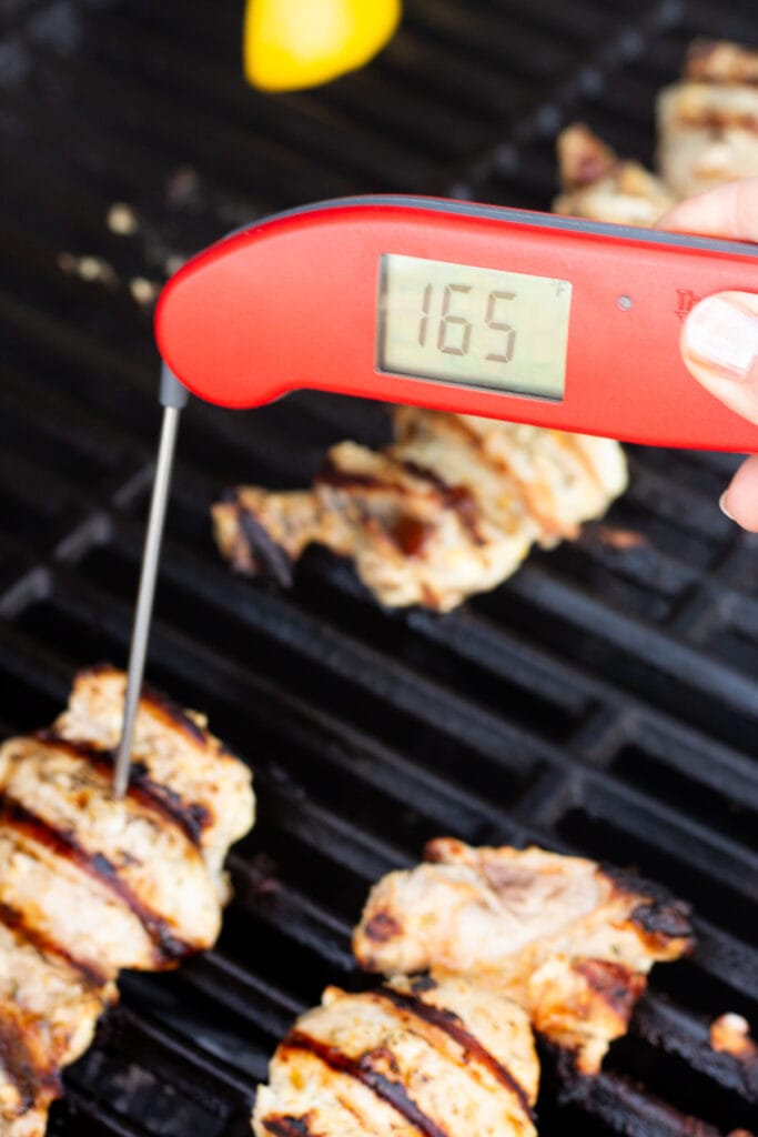 A red meat thermometer measuring the internal temperature of yogurt marinated chicken thighs on a grill, with the temp reading 165F.
