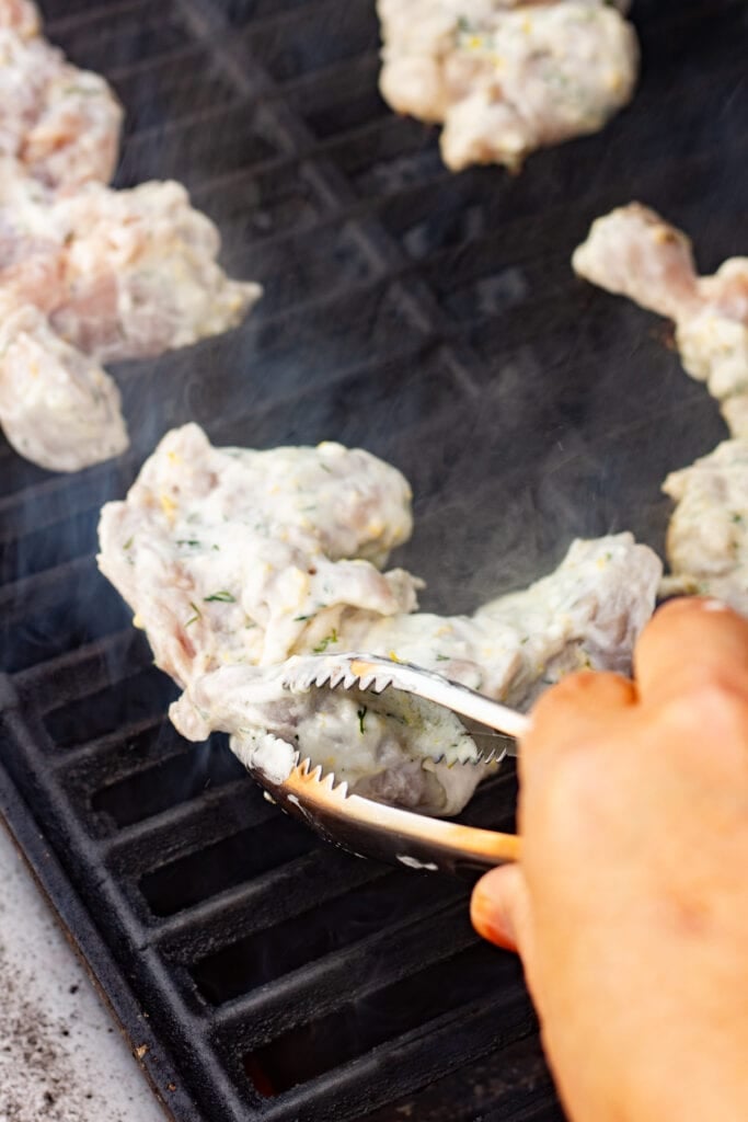 A pair of metal tongs placing yogurt marinated chicken thighs on a hot grill.