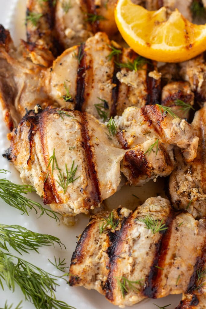 Close up of grilled yogurt-marinated chicken thighs on a white platter with fresh dill and grilled lemon as garnishes.