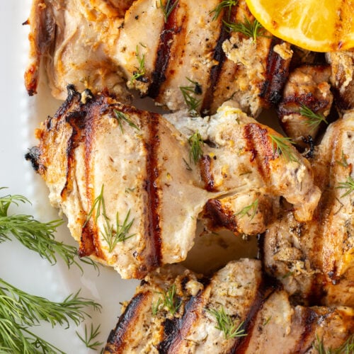 Close up of grilled yogurt-marinated boneless skinless chicken thighs on a white platter with fresh dill and grilled lemon as garnishes.