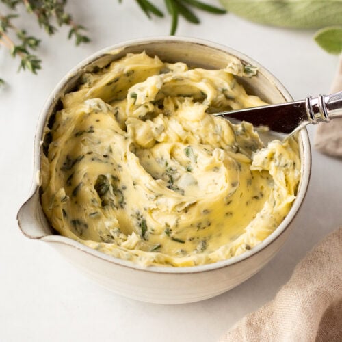 A small bowl with compound herb butter in it, with a butter knife sticking out. The bowl is surrounded by fresh thyme, rosemary, and sage as well as a tan linen napkin.