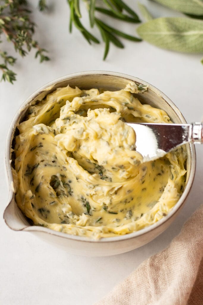 A knife dipping into a small bowl of herb butter that's surrounded by fresh sage, rosemary, and thyme.