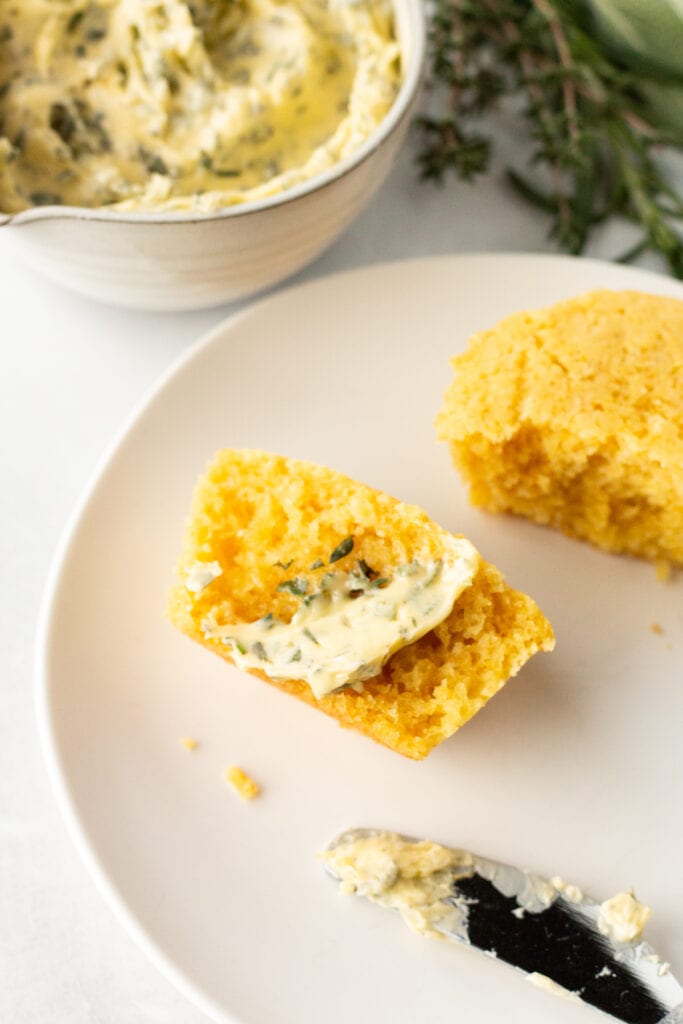 A halved cornbread muffin with a swipe of herb butter on it on a small white plate. A butter knife rests next to it and a small bowl with herb butter, surrounded by fresh herbs, is behind the plate.