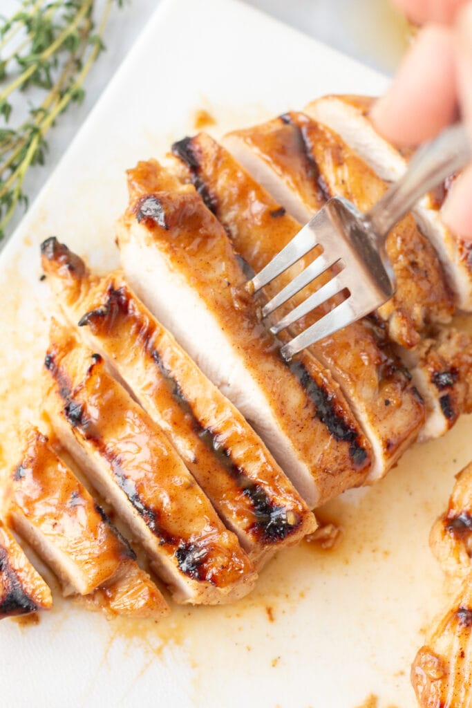 A fork taking a slice of grilled honey mustard chicken off a white cutting board.