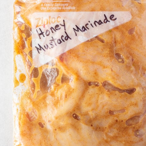 Raw chicken breast covered in a brownish-orange marinade in a gallon sized ziploc bag with the words "honey mustard marinade" written in black sharpie on the front.