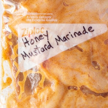 Close up of raw chicken breast covered in a brownish-orange marinade in a gallon sized ziploc bag with the words "honey mustard marinade" written in black sharpie on the front.