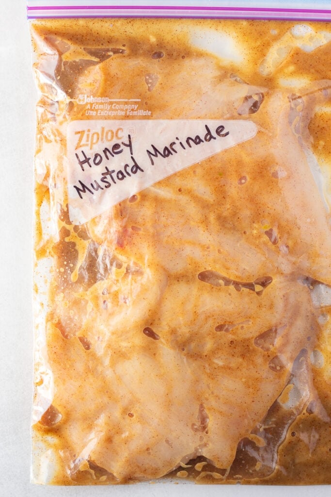 Top down shot of raw chicken breast covered in a brownish-orange marinade in a gallon sized ziploc bag with the words "honey mustard marinade" written in black sharpie on the front.