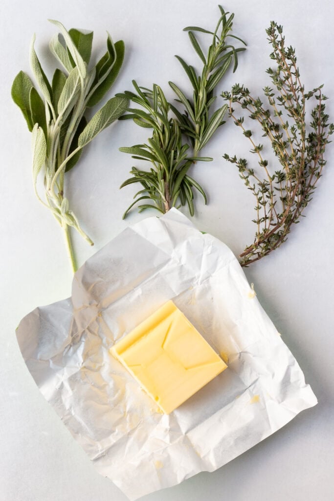 Top down shot of fresh sage, rosemary, and thyme with a slab of butter on a wrapper, all on a white background.