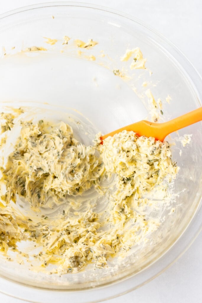 A red spatula scooping out herb butter from a large clear bowl.