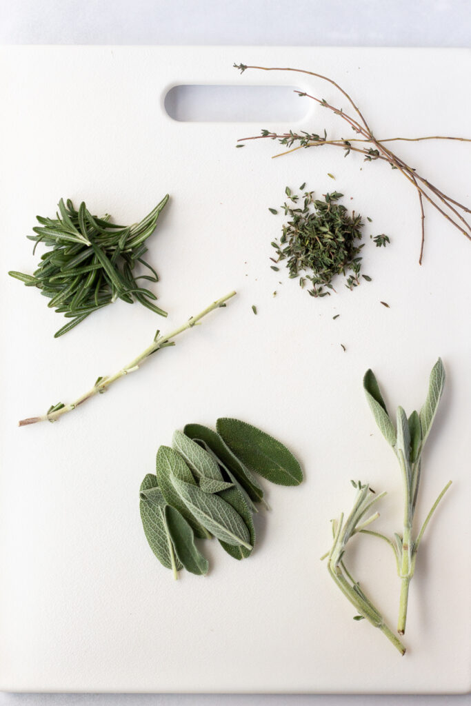 Top down shot of fresh sage, rosemary, and thyme removed from their stems on a white cutting board.