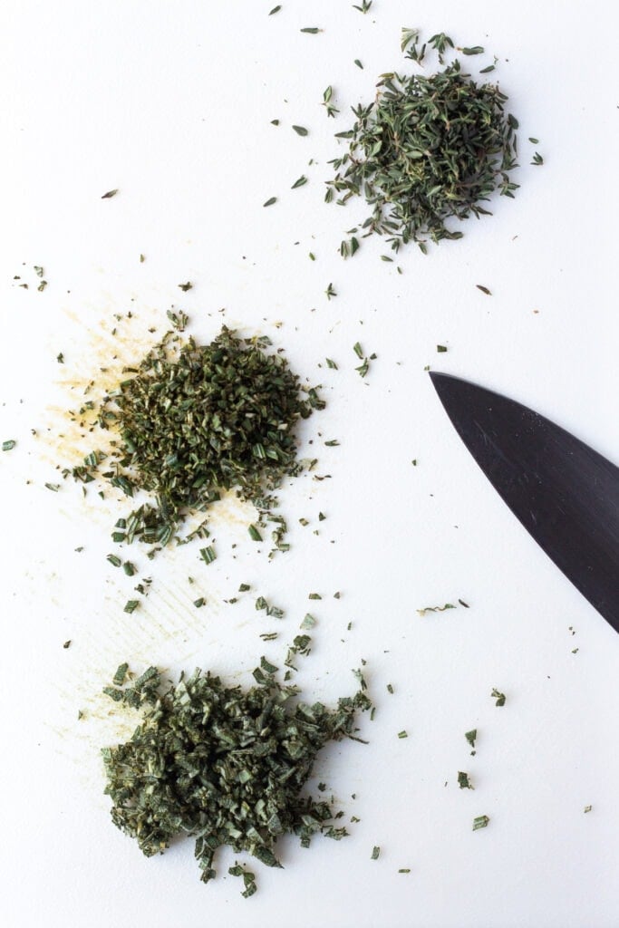 Finely chopped fresh sage, rosemary, and thyme on a white cutting board.