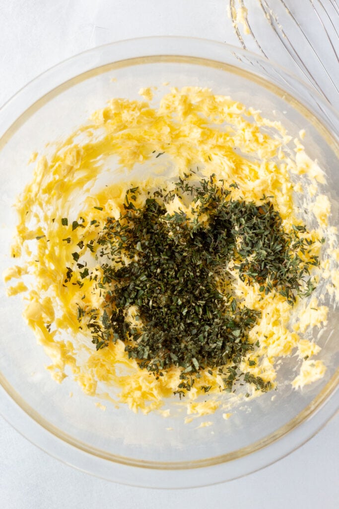 Fresh finely chopped herbs in a large clear bowl with whipped butter in it.