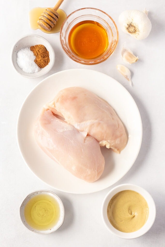 Top down shot of ingredients for grilled honey dijon chicken on a white background, including bowls with honey, mustard, oil, and spices, a plate with two raw boneless chicken breasts, and some fresh garlic.