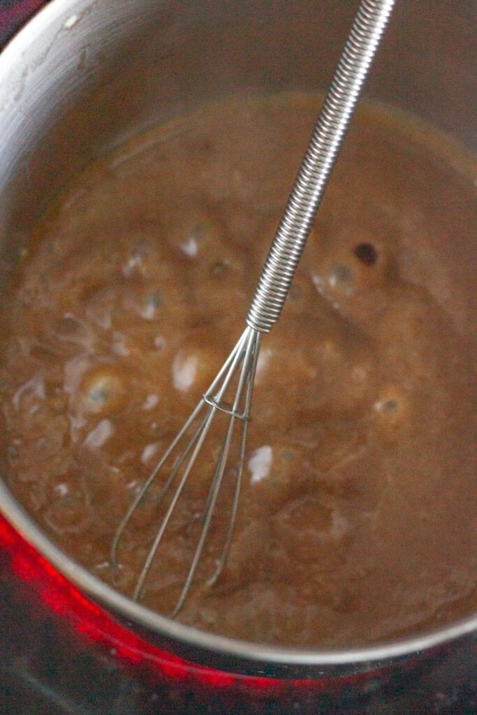 A brown honey mustard glaze boiling in a small sauce pan on the stove. The pan has a small whisk in it as well.