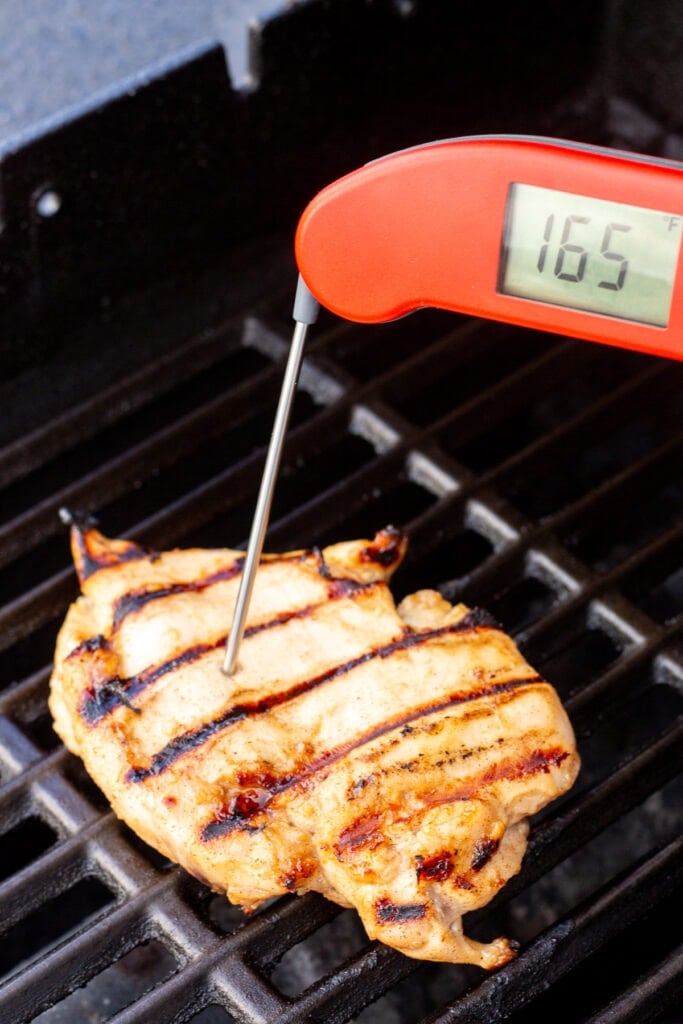 A red meat thermometer being inserted into a grilled chicken breast on a hot grill. The thermometer reads 165F.