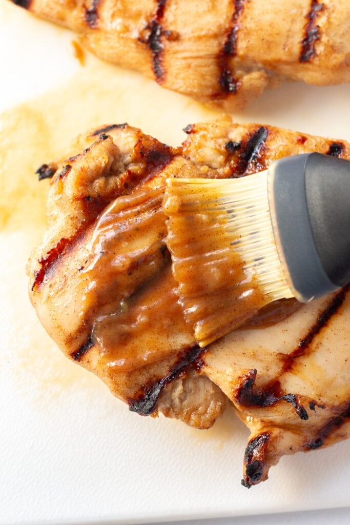A brown glaze being brushed over a grilled chicken breast on a white cutting board.