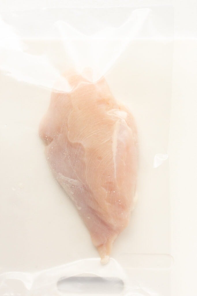 A raw chicken breast in a ziploc plastic bag on a white cutting board.