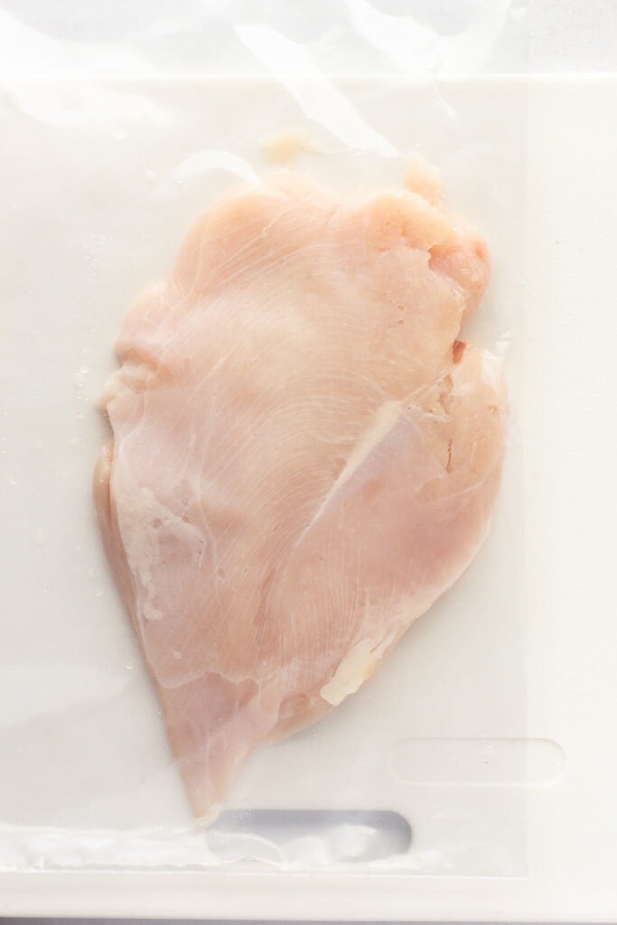 A flattened raw chicken breast in a plastic ziploc bag on a white cutting board.