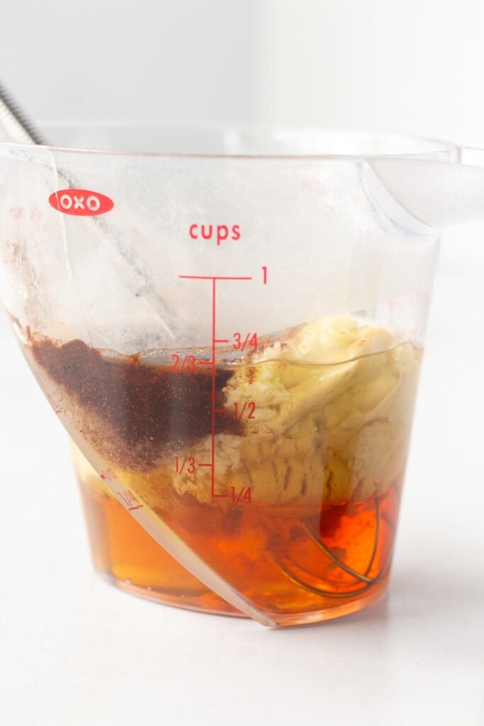 Straight on shot of a liquid measuring cup with honey at the bottom and other ingredients on top, with a small whisk sticking in it as well, all on a white background.