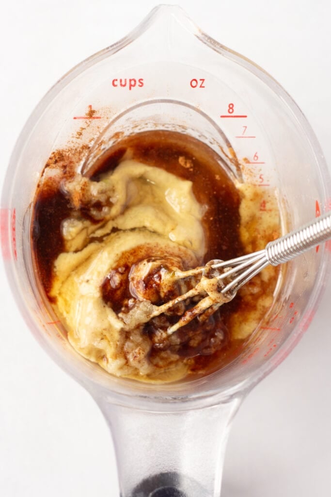 Close up of a small whisk stirring mustard, cinnamon, honey, and other ingredients together in a liquid measuring cup.