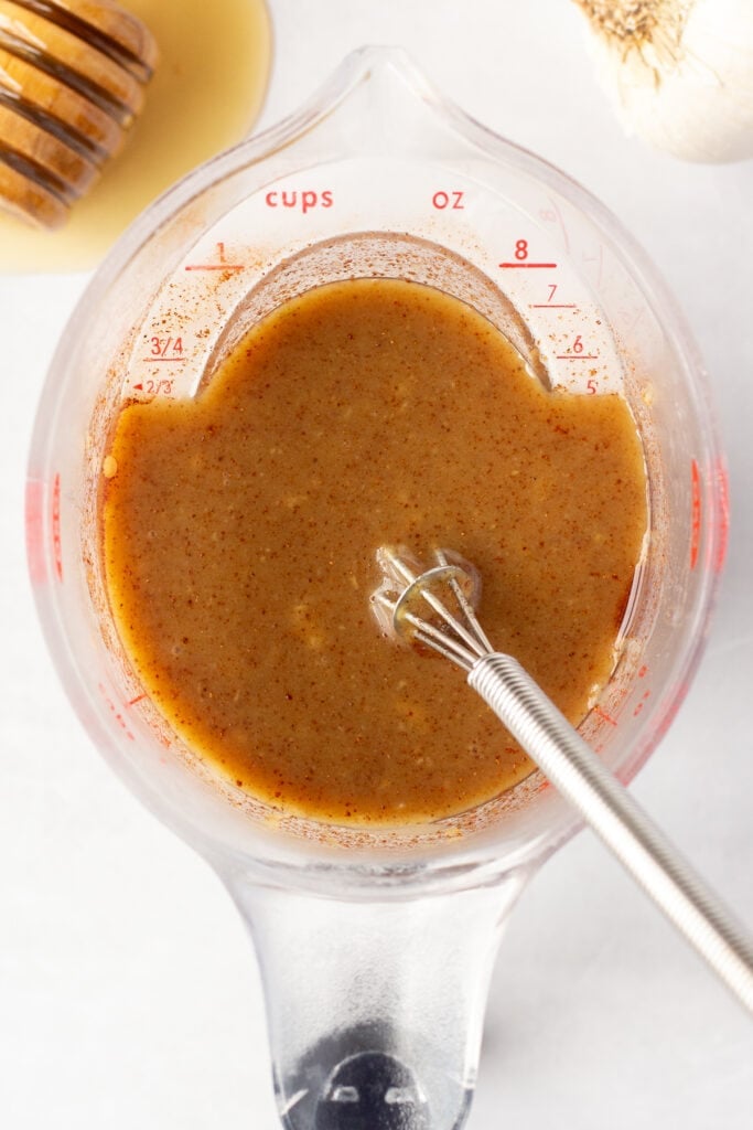 Top down shot of honey mustard marinade in a liquid measuring cup on a white background. A honey stick with honey on it and a head of garlic are next to it.