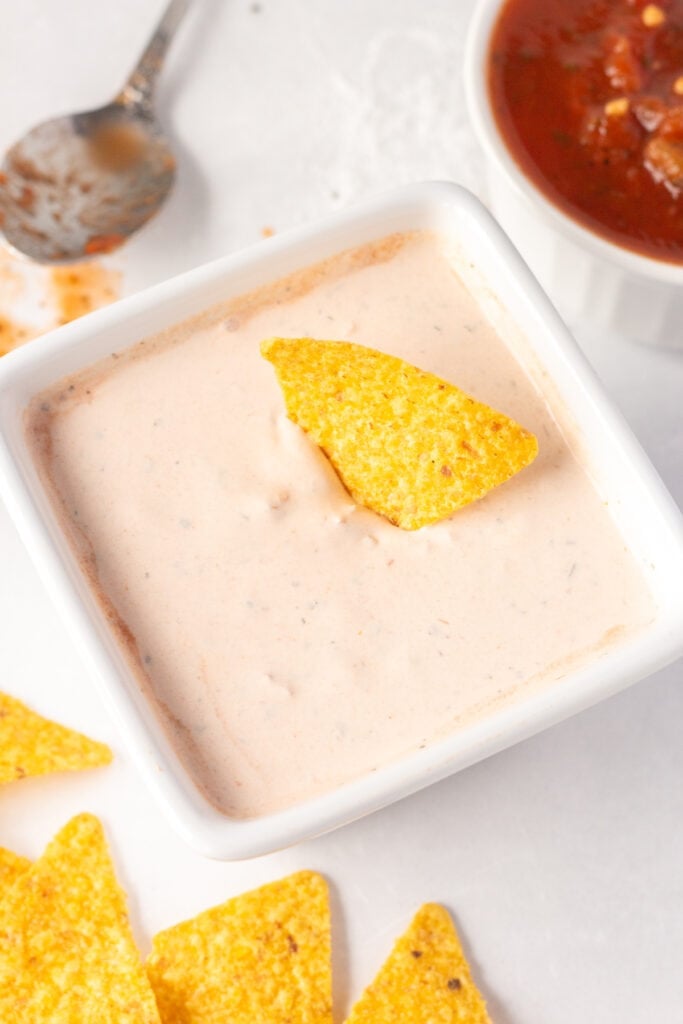 Close up of a white square dish with a salsa sour cream dip in it and a yellow tortilla chip sticking out of it. There is salsa, a spoon, and other tortilla chips surrounding the dish.