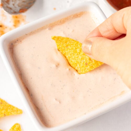 Close up of a hand dipping a yellow tortilla chip into a salsa sour cream dip that's in a white square dish. Surrounding the dish are a bowl of salsa, a dirty spoon with salsa on it, and yellow tortilla chips.